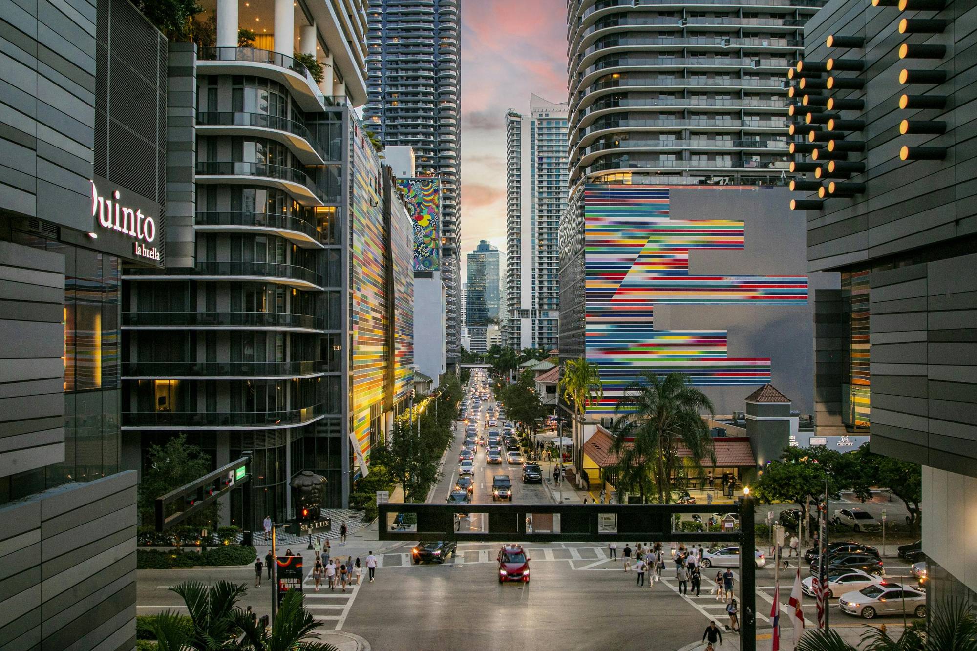 Brickell, Miami at dusk: a street lined with glass towers and painted murals, traffic heading toward the skyline.